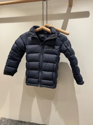 Mörkblå Peak Performance dunjacka 140 - Säljer en mörkblå dunjacka från Peak Performance i storlek 140. Jackan har quiltad design, dragkedja framtill och broderad logga på bröstet. Perfekt nu till vintern. Priset kan diskuteras!