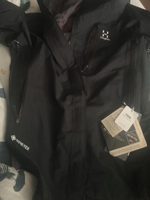 Svart Haglöfs Gore-Tex vindjacka - Säljer en svart vindjacka från Haglöfs i storlek S. Jackan är tillverkad i slitstark Gore-Tex med meshfoder, har flera vattentäta dragkedjor och hög krage. Perfekt för dig som vill ha en funktionell och stilren jacka till vardagsäventyr.