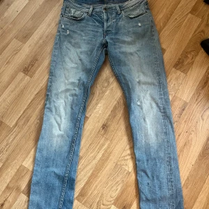 Blå jeans från NN07 model James, storlek 31/32 - Säljer ett par klassiska blå jeans från NN07 i storlek 31/32. Jeansen har en rak passform med slitningar och tvättade detaljer för en avslappnad look. Femficksmodell med knappgylf och snygga kontrastsömmar. Perfekta till sneakers eller boots.