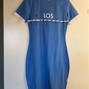 Blå t-shirtklänning Los Angeles FB Sister - Blå t-shirtklänning från FB Sister med vit text 'Los Angeles' över bröstet. Klänningen har korta ärmar med vita muddar och en vit rand över bröstet. Den är figurnära och har en sportig vibe, perfekt för dig som gillar streetstyle.