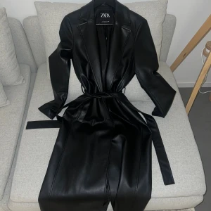 Zara trenchcoat i svart skinn - Säljer en svart trenchcoat från Zara i skinnimitation med bälte i midjan och klassisk krage. Jackan har lång ärm och rak siluett som kan dras åt vid midjan. Perfekt för hösten. 