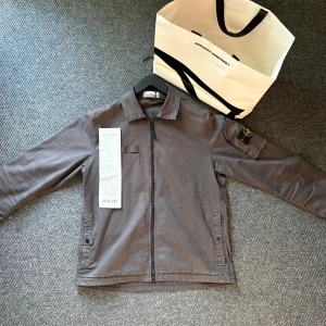 overshirt från Stone Island L - Säljer en stilren grå overshirt från Stone Island i storlek L. Jackan har dragkedja framtill, klassisk krage och två fickor med knappstängning. Ikonisk patch på ärmen och minimalistisk design som passar till många outfits. Jackan är helt ny inköpt på NK i Göteborg med kvitto kvar som medföljer. Aldrig använd super snygg i storlek L men passar M dem som är runt 180 perfekt fit.