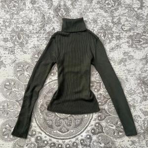 Mörkgrön ribbad polotröja - Säljer en mörkgrön ribbad polotröja med lång ärm och hög krage. Tröjan är figurnära och har en snygg, stretchig passform. Perfekt att styla med jeans eller kjol för en höstig look