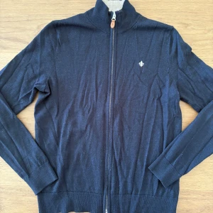 Marinblå kofta från Morris i merinoull - Snygg marinblå kofta från Morris i 100% merinoull. Tröjan har hel dragkedja framtill, ribbade muddar och en liten broderad logga på bröstet. Den är i mycket bra skick och i storlek S.