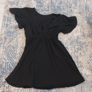 Svart kortklänning med v-ringning - Snygg svart kortklänning med v-ringad hals och dekorativa spetsdetaljer längs ringningen. Klänningen har korta, lite puffiga ärmar och ett mjukt, flowy tyg som ger en skön känsla. Perfekt för dig som gillar enkel men stilren design.