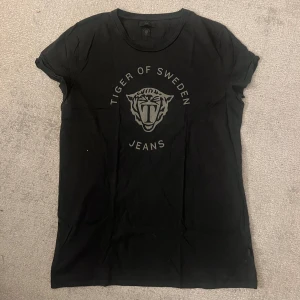 Svart t-shirt Tiger of Sweden Jeans - Svart t-shirt från Tiger of Sweden Jeans med tryckt tigerlogo och text på bröstet. Klassisk rund halsringning och korta ärmar. Skön passform och stilren design, perfekt för dig som gillar streetwear och snygga basplagg. Väligt skön men säljer pågrund av att min bror inte använder den.