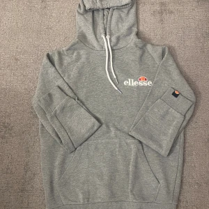 Grå hoodie från Ellesse med logga - Säljer en grå hoodie från Ellesse med vit broderad logga och orange detalj på bröstet. Tröjan har huva med snörning, magficka och ribbade muddar. Perfekt för dig som gillar en sportig och avslappnad stil.