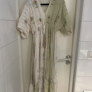 Klänning i vitt och grönt - Superfin långklänning i Positano-stil med v-ringning och korta ärmar. Klänningen är i vitt och ljusgrönt med guldiga broderier och paljettdetaljer, samt knytband i midjan. Materialet är lätt och luftigt. Aldrig använd 