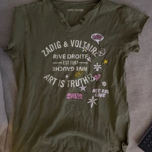 Olivgrön Zadig & Voltaire t-shirt - Olivgrön t-shirt från Zadig & Voltaire med coolt tryck och broderad detalj på ärmen. T-shirten har korta ärmar, v-ringning med knappar och tryck med blommor, stjärnor och text. Tillverkad i mjuk bomull för en skön känsla. I storlek 12 år