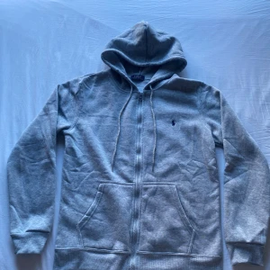 Polo Ralph Lauren kofta  - Snygg grå hoodie från Polo Ralph Lauren med dragkedja och huva. Tröjan har två stora fickor framtill, snörning i huvan och den klassiska broderade loggan på bröstet. Perfekt för chill dagar och enkel att matcha med jeans eller joggers.
