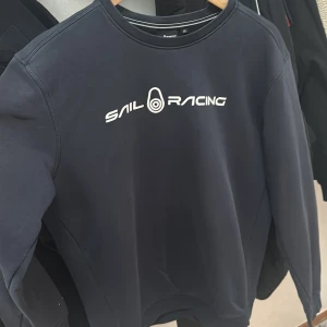 Mörkblå Sail Racing sweatshirt XL - Mörkblå sweatshirt från Sail Racing i storlek XL. Tröjan har rund hals, lång ärm och vit logotyp framtill samt en mindre logga bak i nacken. Tillverkad i mjuk bomullsblandning med ribbade muddar vid ärmslut och nederkant. Perfekt för en chill och sportig look.