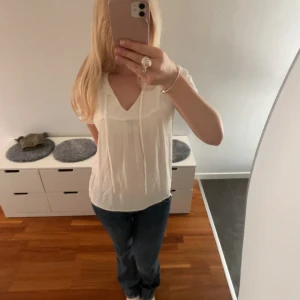 Vit blus med knyt och volang från JDY - Söt vit blus från JDY med korta ärmar, v-ringning och knytband framtill. Blusen har diskreta prickar i tyget, volangdetalj vid halsen och ett luftigt, lätt material. (Priset kan diskuteras)