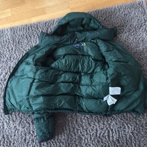 Grön dunjacka från Polo Ralph Lauren - Mörkgrön pufferjacka från Polo Ralph Lauren med huva och dragkedja framtill. Jackan har två fickor med dragkedja och är vadderad för extra värme. Perfekt för kyliga dagar när du vill hålla stilen.