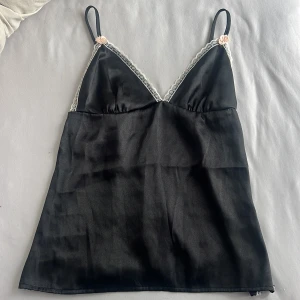 Svart satin linnetopp med spets och rosetter - Supersöt svart linnetopp i satin med justerbara tunna axelband, vit spetskant och små ljusrosa rosetter framtill och nertill. Toppen har v-ringning och smock i ryggen för extra komfort. Perfekt för en trendig och 90s look.