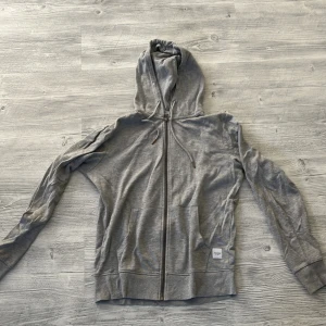 Grå hoodie med dragkedja Jack & Jones - Grå hoodie från Jack & Jones med hel dragkedja framtill, justerbar huva med snören och två fickor. Klassisk och enkel design med liten logolapp på nedre delen. Perfekt för chill dagar eller när du vill ha något bekvämt.