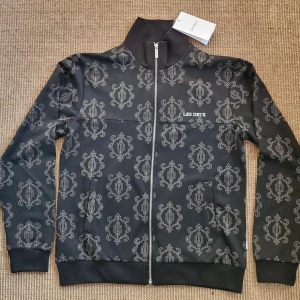 Svart mönstrad tjock tröja från Les Deux  - Svart tjock tröja från Les Deux med hel dragkedja och hög krage. Jackan har ett stilrent, grått mönster över hela ytan och ribbade muddar vid ärmslut och nederkant. Perfekt för dig som vill sticka ut med en snygg streetstyle. Säljer tjock tröjan pågrund av att jag fick den i fölsedagspresent och kunde inte lämna tillbaka den, för att den blev köpt i Danmark. Helt ny rakt ur affären.