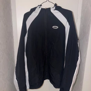 Cortiez jacka - Svart vindjacka med vita paneler längs ärmar och axlar. Jackan har huva, hel dragkedja framtill och sportig look. Liten logga på bröstet. Perfekt för dig som gillar streetwear och vill ha en lätt jacka till vardags.