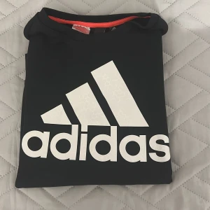 Svart Adidas sweatshirt med logga - Svart långärmad sweatshirt från Adidas med stor vit logga framtill och gråa detaljer på ärmarna. Tröjan har rund halsringning, dragkedjor vid sidorna och är tillverkad i ett mjukt syntetmaterial. Perfekt för träning eller chill. Pris kan diskuteras!