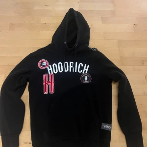 Svart hoodie från Hoodrich med patchar - Svart hoodie från Hoodrich med stor huva och coola broderade patchar och tryck framtill. Tröjan har snörning i huvan, känguruficka och ribbade muddar. Perfekt för dig som gillar streetwear och vill sticka ut med snygga detaljer.