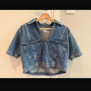Croppad jeansblus från Zara - Säljer en croppad jeansblus från Zara i klassisk blå denim. Modellen har korta ärmar, två stora bröstfickor, krage och v-ringning med dragkedja framtill. Perfekt till ett par höga byxor för en cool look.