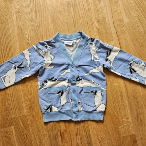 Blå kofta med kaninmönster Mini Rodini - Superfin blå kofta från Mini Rodini med sött kaninmönster i vitt och svart. Koftan har ribbade muddar, två fickor framtill och stängs med blå knappar. Perfekt för dig som gillar lekfull stil och vill sticka ut lite extra.