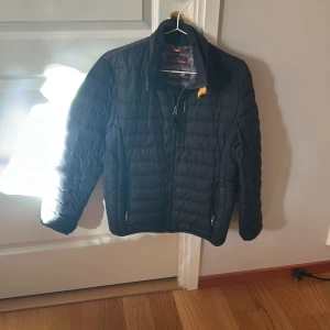 Svart lättviktsjacka Parajumpers Y-M - Säljer en svart superlätt pufferjacka från Parajumpers i storlek Y-M. Jackan har quiltad design, dragkedja framtill och två sidofickor med dragkedja. Klassisk ståkrage och orange detalj vid halsen. Perfekt för dig som vill ha en snygg och smidig jacka.