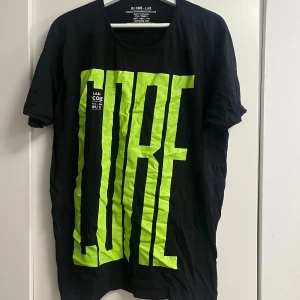 Svart t-shirt med neongrön print CORE - Cool svart t-shirt från J&J CORE - L.A.B. med stor neongrön text 'CORE' framtill. Klassisk rund hals och korta ärmar. Skön bomullskvalitet som passar perfekt till jeans eller shorts. Snygg streetstil med tydlig logga och tryckdetaljer.