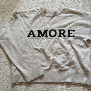 Beige stickad tröja med AMORE-print - Beige stickad tröja från Gina Tricot med svart AMORE-tryck över bröstet. Tröjan har rund halsringning, ribbade muddar och lång ärm. Perfekt för dig som gillar en enkel men snygg look.