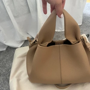 Beige handväska från Polene i skinn! Super snygg!! Köpt begagnat men inte mycket använd av någon ägare! Prislappen sitter på och detta är den stora sizen! 💗💗 - Snygg beige handväska från Yuzefi i mjukt, strukturerat skinn. Väskan har en unik form med dubbla handtag, guldfärgade metalldetaljer och justerbar axelrem. Perfekt för dig som vill ha en stilren och trendig accessoar med plats för det viktigaste.