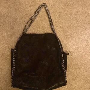 Svart axelväska med kedjerem - Cool svart axelväska med glansig yta och grova silverfärgade kedjor som axelrem. Väskan har en mjuk, slouchy form och är perfekt för att rymma allt du behöver. Kedjedetaljerna ger en edgy vibe och väskan passar till många olika outfits.