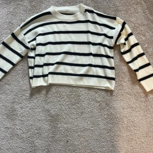 Randig stickad tröja i vitt och svart - Snygg stickad tröja med svarta ränder på vit botten. Tröjan har en avslappnad passform, rund halsringning och långa ärmar. Perfekt för lager-på-lager och passar till både jeans och kjol.