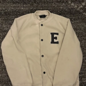 Beige bomberjacka från Everest - Säljer en beige bomberjacka från Everest med svart E-patch på bröstet. Jackan har ribbade muddar, knappar framtill och är tillverkad i mjukt syntetmaterial. Perfekt för dig som vill ha en clean och stilren look.