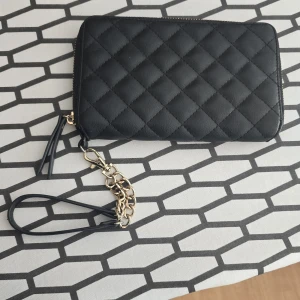 Svart quiltad plånbok Don Donna - Stilren svart quiltad plånbok från Don Donna i skinnimitation med guldfärgad kedjedetalj och dragkedja runtom. Flera kortfack, myntfack med dragkedja och avtagbar handledsrem. 