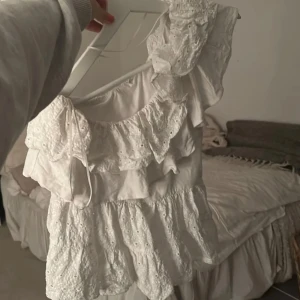 Vit volangblus med spetsdetaljer - Supersöt vit blus med flera lager volanger och broderade spetsdetaljer. Blusen har en romantisk känsla med omlottkänsla och knytband vid axeln. Perfekt för dig som gillar en feminin och luftig stil.