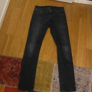 Svarta jeans från Nudie Jeans - Säljer ett par svarta jeans från Nudie Jeans med klassisk femficksdesign och snygga detaljer på bakfickorna. Jeansen har en rak passform och är tillverkade i slitstarkt denimtyg. Perfekta för dig som gillar stilrena och tidlösa jeans.