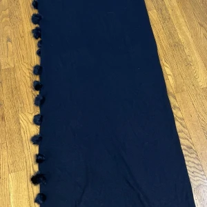 Svart halsduk med tofsar - En lång svart halsduk med fluffiga tofsar längs kanterna och fransar i ena änden. Halsduken är mjuk och har en enkel, stilren look som passar perfekt för kalla dagar. Materialet känns som stickad syntet eller ullblandning.