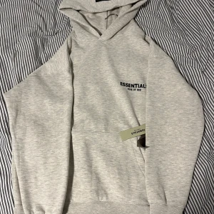 Grå Essentials Fear of God hoodie S - Snygg ljusgrå hoodie från Essentials Fear of God med svart logga på bröstet, luvan och ryggen. Klassisk känguruficka framtill. Mjuk insida i bomullsblandning, perfekt för höst och vinter. 