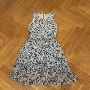Blommig blåvit ärmlös sommarklänning - Superfin ärmlös klänning med blått blommönster på vit botten. Klänningen har volang nertill och markerad midja för en snygg siluett. Perfekt för varma dagar och har en lätt, luftig känsla.