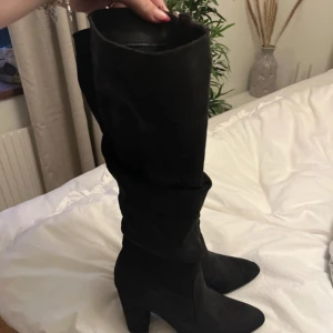 Svarta höga stövlar med klack - Säljer ett par svarta höga stövlar i mocka-look med chunky klack och spetsig tå. Stövlarna har en stilren design och går upp över knäet, perfekta till både kjol och jeans. Materialet är mjukt och ger en lyxig känsla. Använda en gång säljer då de ej passar längre 