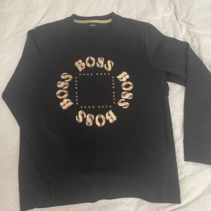 Svart sweatshirt från Hugo Boss - Svart sweatshirt från Hugo Boss med rund hals och tryck i guld och beige på bröstet där det står BOSS i en cirkel. Sparsamt använt! Storlek S. Inga skador eller liknande!