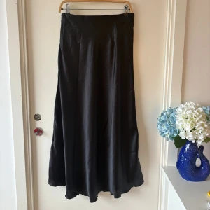 Svart långkjol i satin  - Säljer en svart långkjol från Angelology i glansigt satinliknande material. Kjolen har en stilren, lätt utställd passform och dold dragkedja i sidan. Perfekt för dig som gillar en clean och elegant look. Stretchig i midjan   Endast testad, aldrig använd 