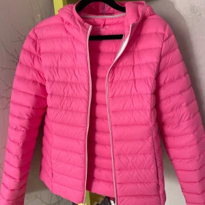 Rosa dunjacka från Marie Lund - Snygg rosa dunjacka från Marie Lund med huva och horisontella sömmar. Jackan är lätt och har dragkedja framtill. Perfekt för dig som vill sticka ut med en färgstark look under kyliga dagar. Strl 38.