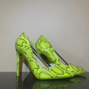 Neongula pumps med ormmönster - Säljer ett par riktigt snygga pumps i neongult med ormmönster. Skorna har spetsig tå och hög klack, vilket ger en edgy vibe. Materialet är glansigt syntet som verkligen sticker ut. Perfekta för dig som vill synas och älskar statement pieces.