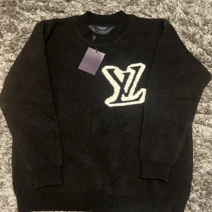 Svart stickad tröja från Louis Vuitton - Svart stickad tröja från Louis Vuitton med stor vit LV-logga på bröstet. Tröjan har rund halsringning, ribbade muddar och är långärmad. Perfekt för dig som vill ha en lyxig och stilren look med streetkänsla.