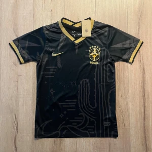 Svart Brasil Nike fotbollströja S - Snygg svart Brasilien fotbollströja från Nike med guldiga detaljer och broderat CBF-märke på bröstet. Tröjan har V-ringning, korta ärmar och är tillverkad i Dri-FIT-material som andas. Unika grafiska mönster ton-i-ton på framsidan och guldiga kanter vid hals och ärmslut.