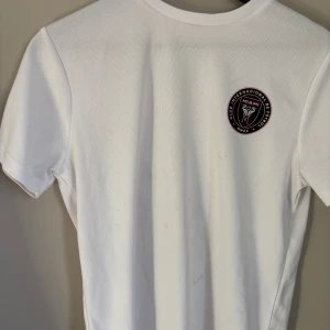 Vit Inter Miami fotbollströja MLS - Snygg vit fotbollströja från Inter Miami med klubbens stora logga i svart och rosa på ryggen och mindre logga på bröstet. Tröjan har korta ärmar och är tillverkad i ett lätt, ventilerande material som passar perfekt för träning eller match.