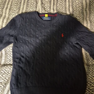 Mörkblå kabelstickad tröja Polo Ralph Lauren - Kabelstickad mörkblå tröja från Polo Ralph Lauren med klassisk röd broderad logga på bröstet. Tröjan har rund halsringning, ribbade muddar och är långärmad. Perfekt för dig som gillar stilren och tidlös design.