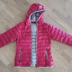 Rosa pufferjacka från Sail Racing S - Snygg rosa pufferjacka från Sail Racing i storlek S. Jackan har huva, två dragkedjeförsedda fickor och grått innerfoder. Perfekt för kyliga dagar och har en sportig look med logga på bröstet och huvan.