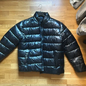 Svart pufferjacka  - Säljer en svart pufferjacka från Livergy i storlek L. Jackan har glansig finish, horisontella sömmar och dragkedja framtill.  Helt ny aldrig använt 