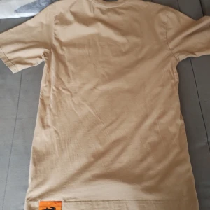 Beige t-shirt från Pacemaker med ficka - Tvär schysst beige t-shirt från Pacemaker med klassisk passform. T-shirten har en bröstficka med svart kanin-tryck som ger en unik touch. Tillverkad i mjuk bomull och har rund halsringning och korta ärmar. Perfekt för en chill och stilren look.